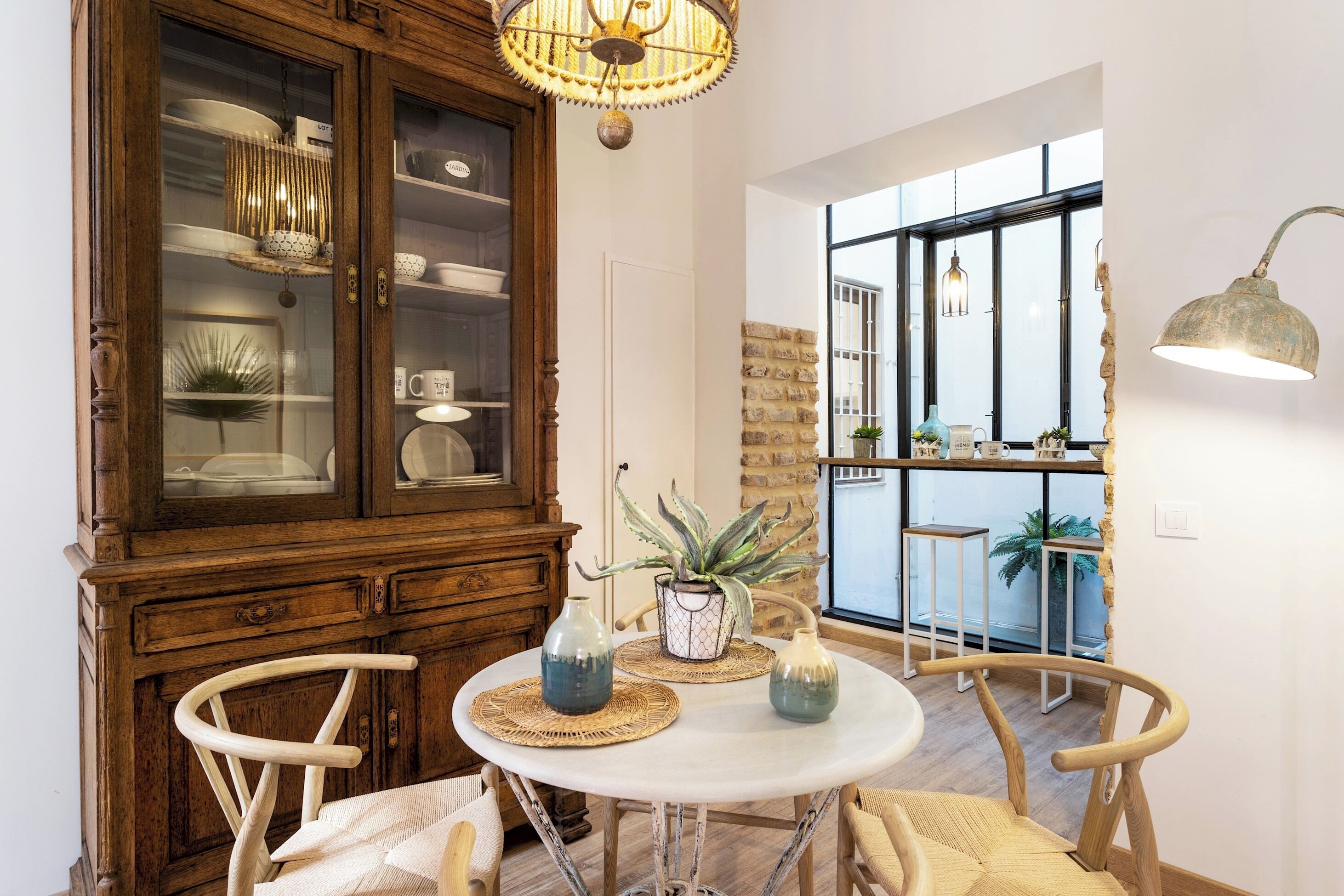 A dining area with a round table, wooden chairs, a cabinet with glass doors, and a view through a large window.