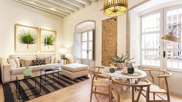 A modern living room with a sofa, ottoman, coffee table, and dining area with chairs. There is a chandelier, a plant, and a view of the outside through large windows.
