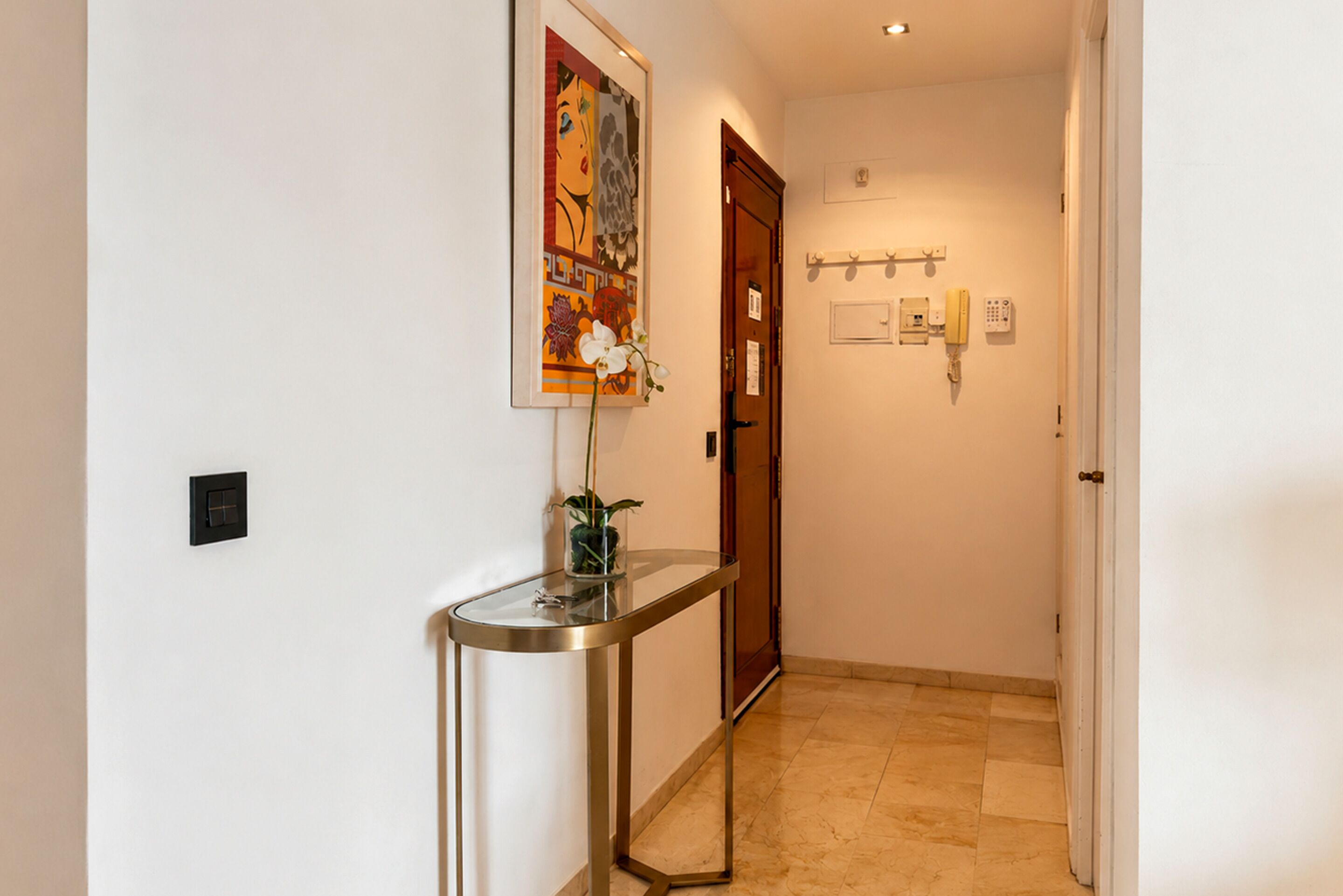 Un pasillo con una recepción, una obra de arte enmarcada, una planta en maceta, una puerta y un teléfono montado en la pared.