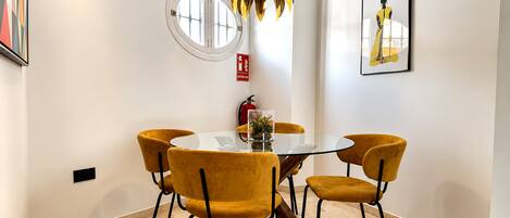 A dining area with a round table, four chairs, a pendant light, a fire extinguisher, and a framed artwork on the wall.