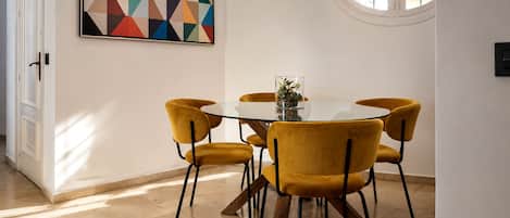 Un comedor con una mesa redonda, cuatro sillas, un cuadro geométrico, una lámpara colgante y una ventana.