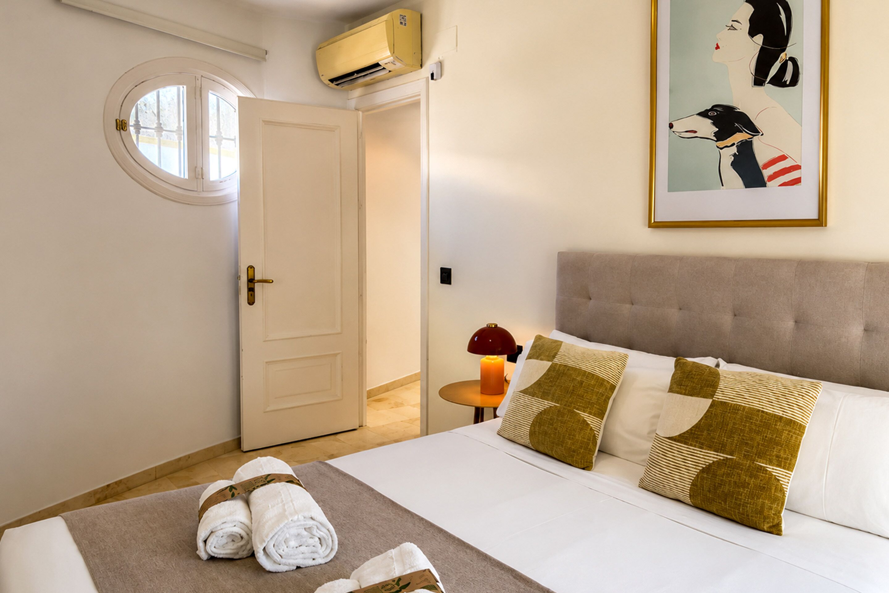 A hotel room with a bed, a door, an air conditioning unit, a framed picture, and a bedside table with a candle.