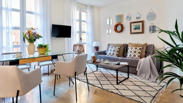 A modern living room with a sofa, coffee table, and chairs, a TV mounted on the wall, and a dining area with a glass table and chairs. - Genteel Home Reyes Católicos (Granada)
