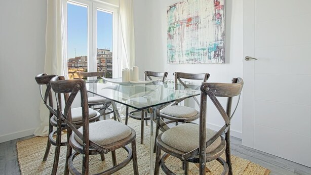 A dining area with a glass table surrounded by wooden chairs, a large abstract painting on the wall, and a view of buildings through the window. - The Best Location, 8pax, 3BD & 2Bth, Reina Sofia II (Madrid)