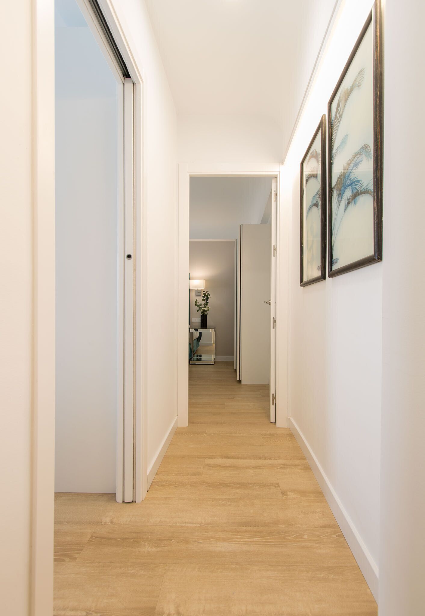 A hallway with wooden flooring and white walls, leading to a room with a bed and a nightstand.