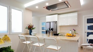 A modern kitchen with white cabinets, a stainless steel refrigerator, and a bar area with white chairs. - Genteel Home Recogidas (Granada)