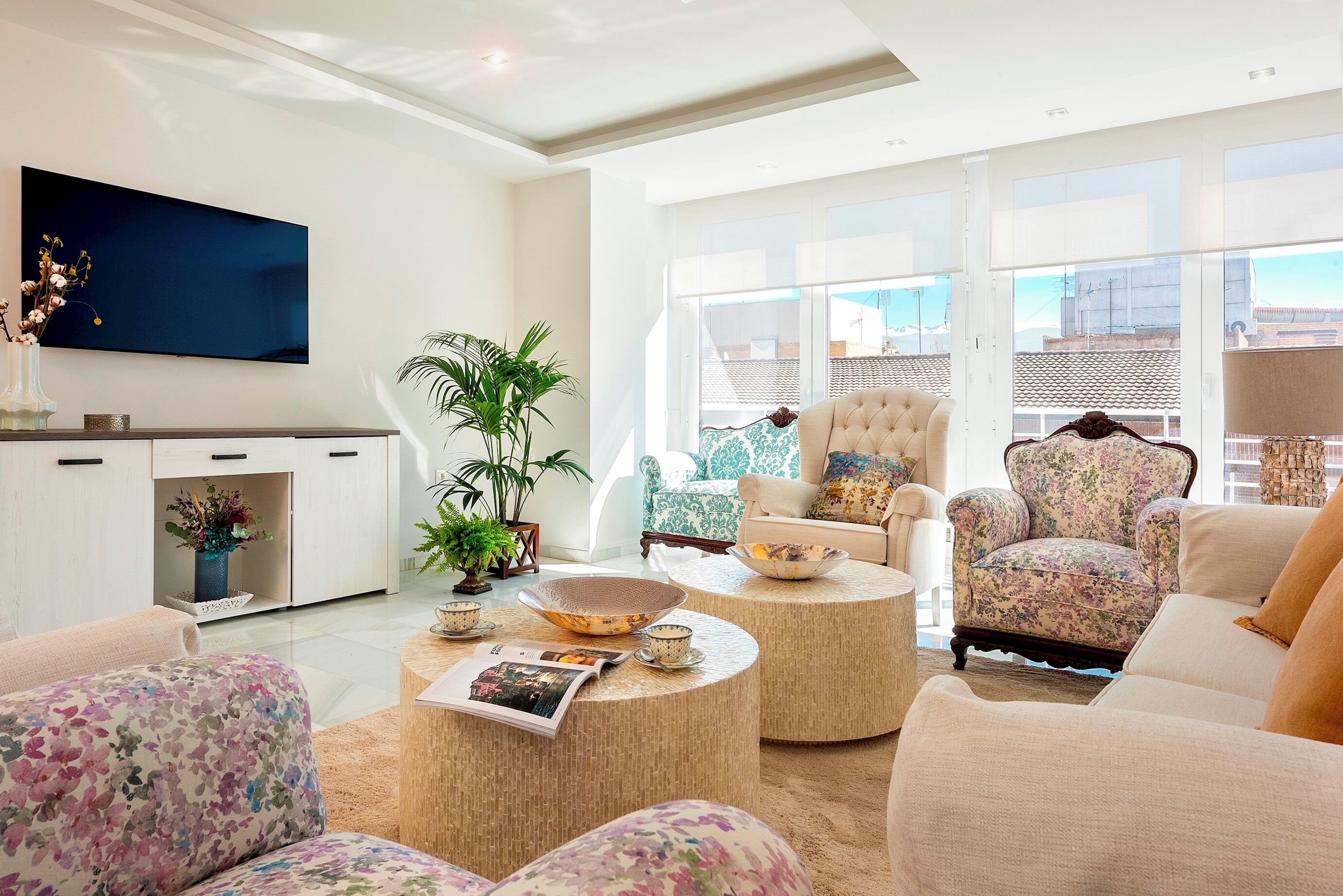 Ein geräumiges Wohnzimmer mit Flachbildfernseher, floral gemusterten Sofas und einem großen Fenster mit Blick auf die Stadt.