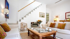 Un salón con una escalera, sofás blancos, una mesa de centro de madera y una mesa con cubierta de vidrio, tazas y un florero.