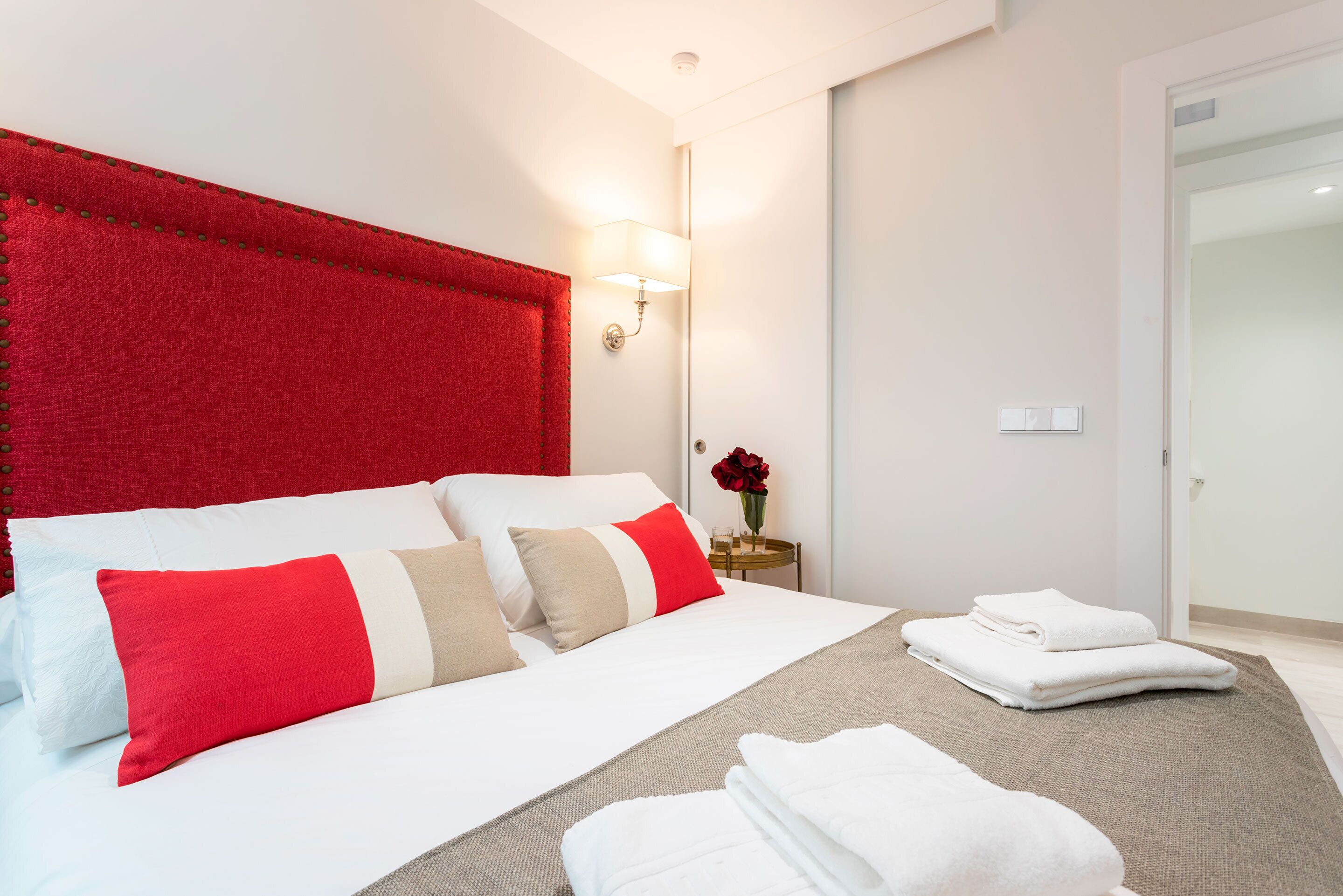 A neatly made bed with white and red bedding, a red headboard, and a bedside table with a vase of red flowers.