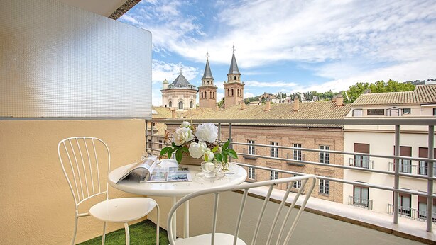 A balcony with a table and chairs, a vase with flowers, and a view of a church with two towers. - Genteel Home Amira (Granada)