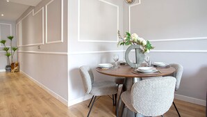 A dining area with a round table, grey upholstered chairs, and a vase with flowers. - Genteel Home Amira (Granada)
