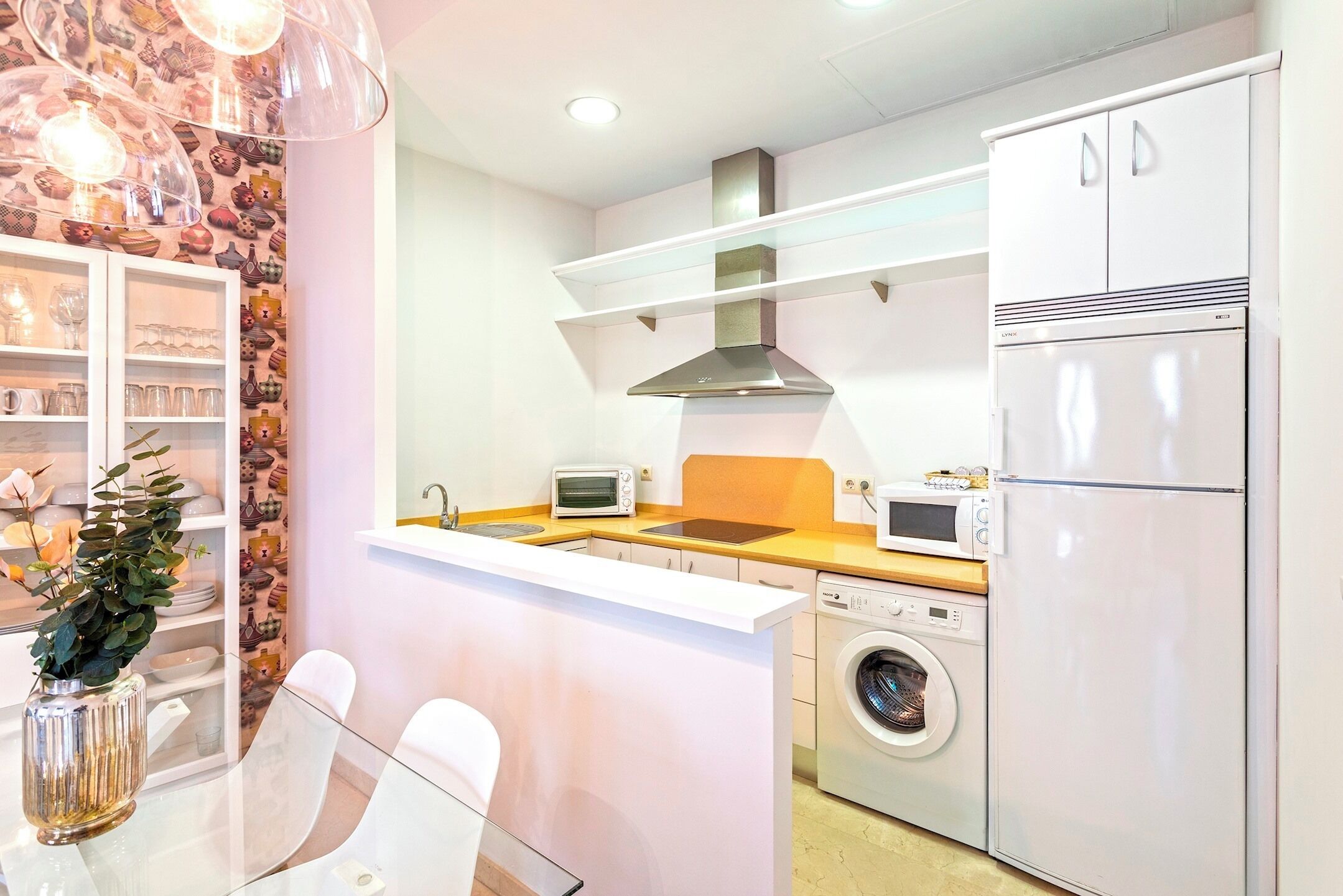 A modern kitchen with a built-in oven, microwave, and refrigerator, a washing machine, and a glass-fronted cabinet with dishes.