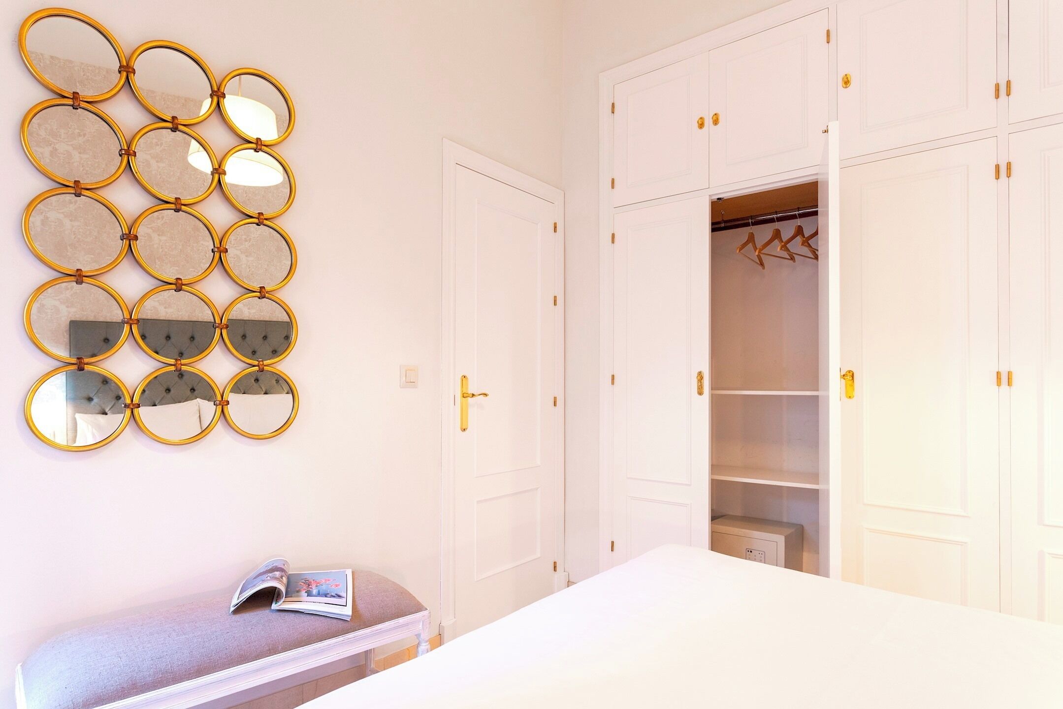 Een slaapkamer met een bed, een nachtkastje met een tijdschrift, een wandspiegel en een garderobe met meerdere deuren en kledinghangers.