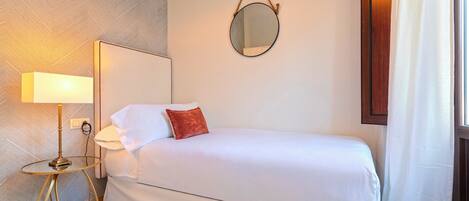 A hotel room with a bed, a bedside lamp, a round mirror, and a window with curtains.