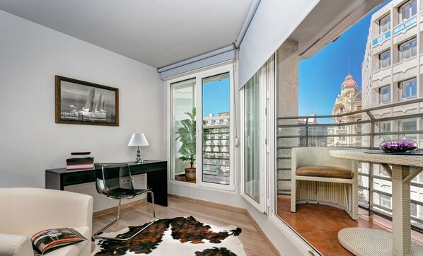 Un salon moderne doté d’un balcon en verre, d’un bureau, d’une chaise, d’un canapé et offrant une vue sur le paysage urbain.