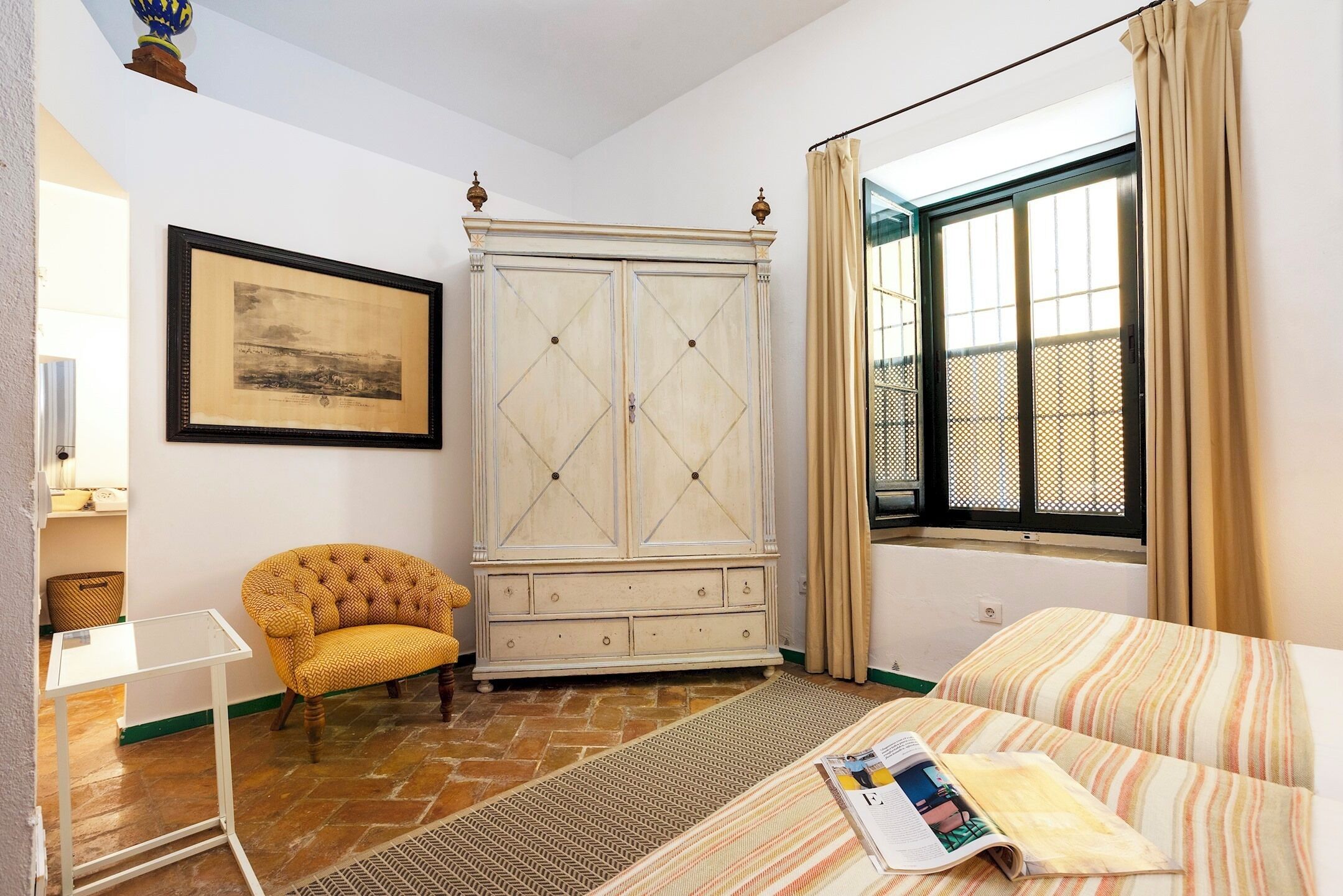 A bedroom with a bed, a wooden wardrobe, a yellow armchair, a framed picture on the wall, and a window with curtains.