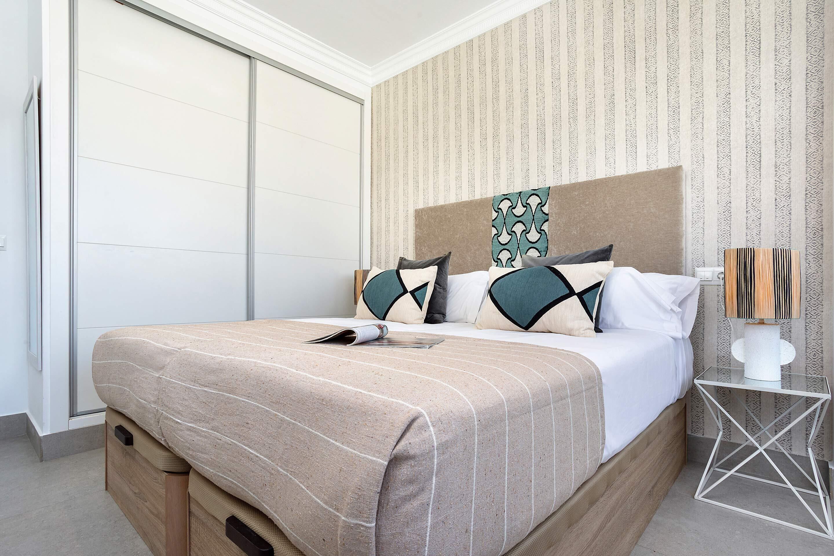 A neatly made bed with a striped bedspread, a headboard with a geometric pattern, and a bedside table with a lamp.