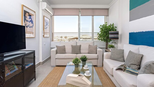 A modern living room with a sofa, coffee table, and a TV. There are framed artworks on the wall, a plant, and a view through large windows. - Genteel Home Campana (Seville)