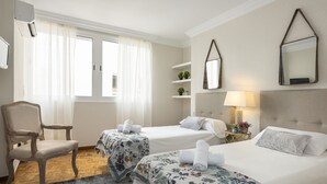 A hotel room with two beds, a chair, a small table with a lamp, and a window with curtains.