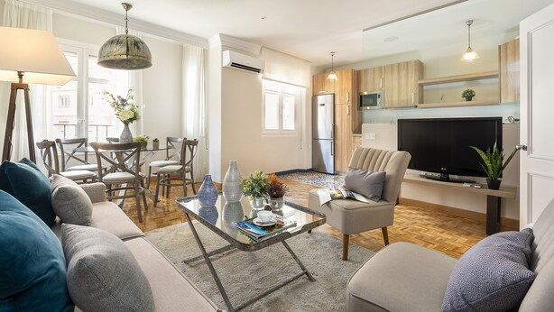A modern living room with a sofa, coffee table, and TV. There is a dining area with chairs and a table. The kitchen is visible with wooden cabinets and a TV.