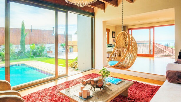 A modern living room with a large glass door leading to a pool, a hanging chair, and a wooden coffee table with decorative items. - Sun & Sea Family Villa (Cape Town)