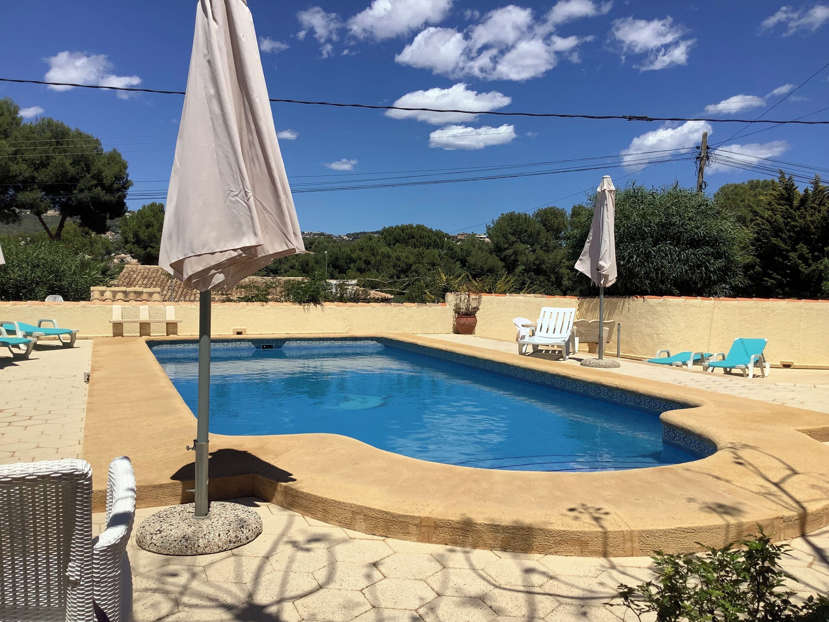 A swimming pool with lounge chairs and umbrellas, surrounded by a landscaped area with trees and a view of the hills.