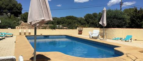 A swimming pool with lounge chairs and umbrellas, surrounded by a landscaped area with trees and a view of the hills.