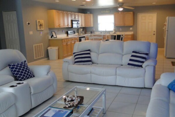 A living room with a white sofa, a glass coffee table, and a kitchen in the background.