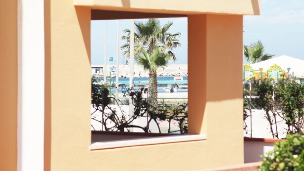A view through a window of a building with a palm tree and a beach in the background. - I tre Golfi Faro Blu (San Vito Lo Capo)