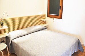 A small, neatly arranged bedroom with a single bed, a bedside table, a chair, and a window with a wooden frame.