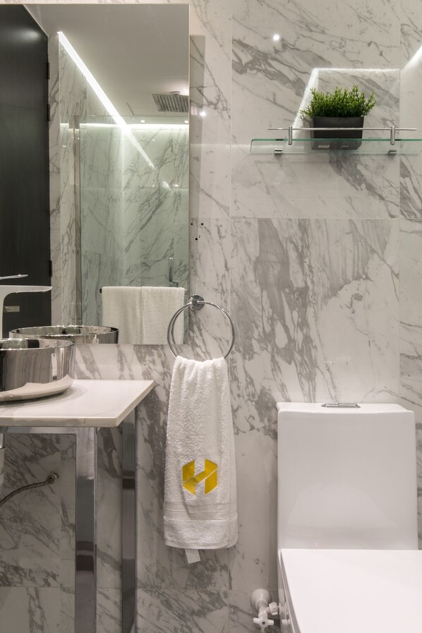 A modern bathroom with a marble backsplash, a glass shower enclosure, a white sink with a stainless steel basin, a towel rack with a white towel, and a small potted plant on a shelf.