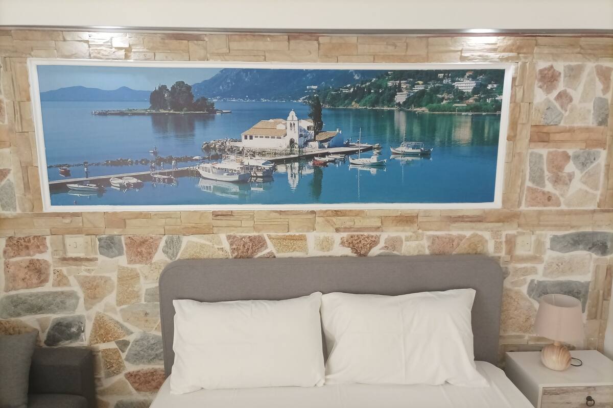 A bedroom with a stone wall, a bed with white bedding, a nightstand with a lamp, and a painting of a harbor scene.