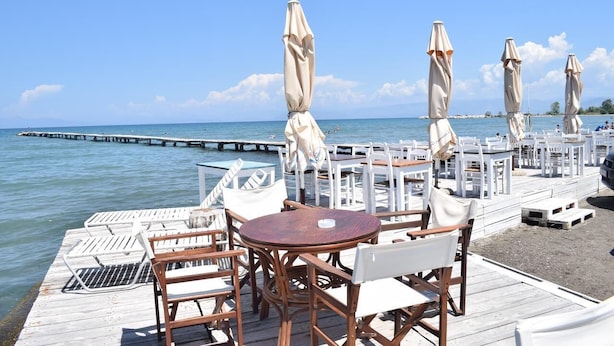 A wooden deck with tables and chairs, a pier extending into the water, and a clear sky. - Petrakis Beach (Corfu)