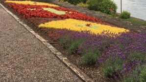 Un jardin avec des parterres de fleurs rouges, jaunes et violettes, situé à côté d’une étendue d’eau.