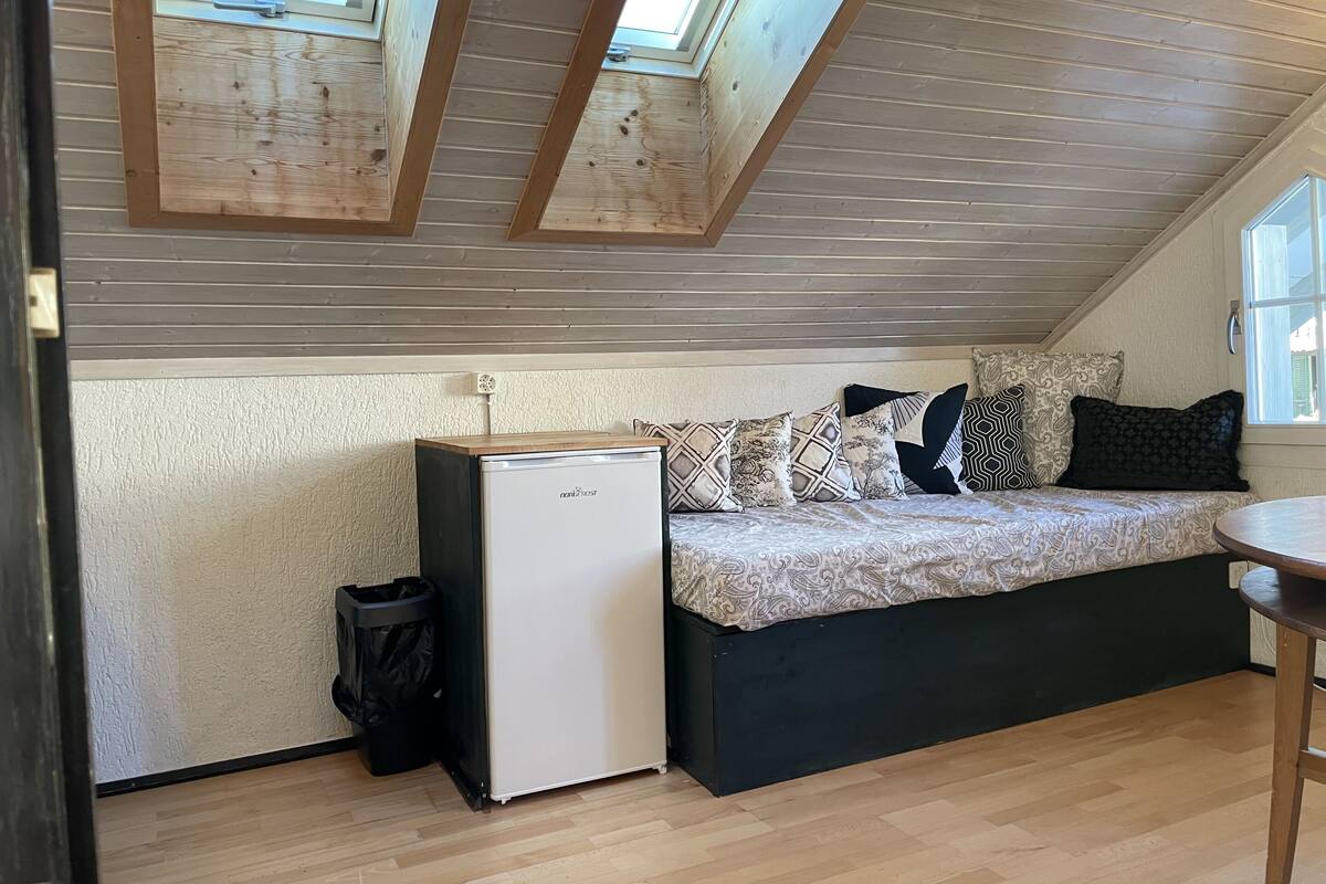 A small, cozy room with a sloped ceiling, a bed with patterned bedding, a small refrigerator, and a window.