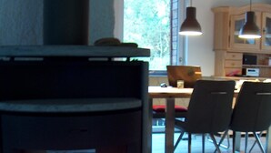 A wood-burning stove with a fire, a dining table with chairs, and a kitchen area with cabinets and pendant lights. - Yellow house: secluded location, sauna hut, terrace, fenced - vacation with dog (Bodendorf)