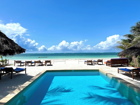 Vista da piscina com água azul cristalina, cadeiras de praia e uma estrutura com telhado de palha.