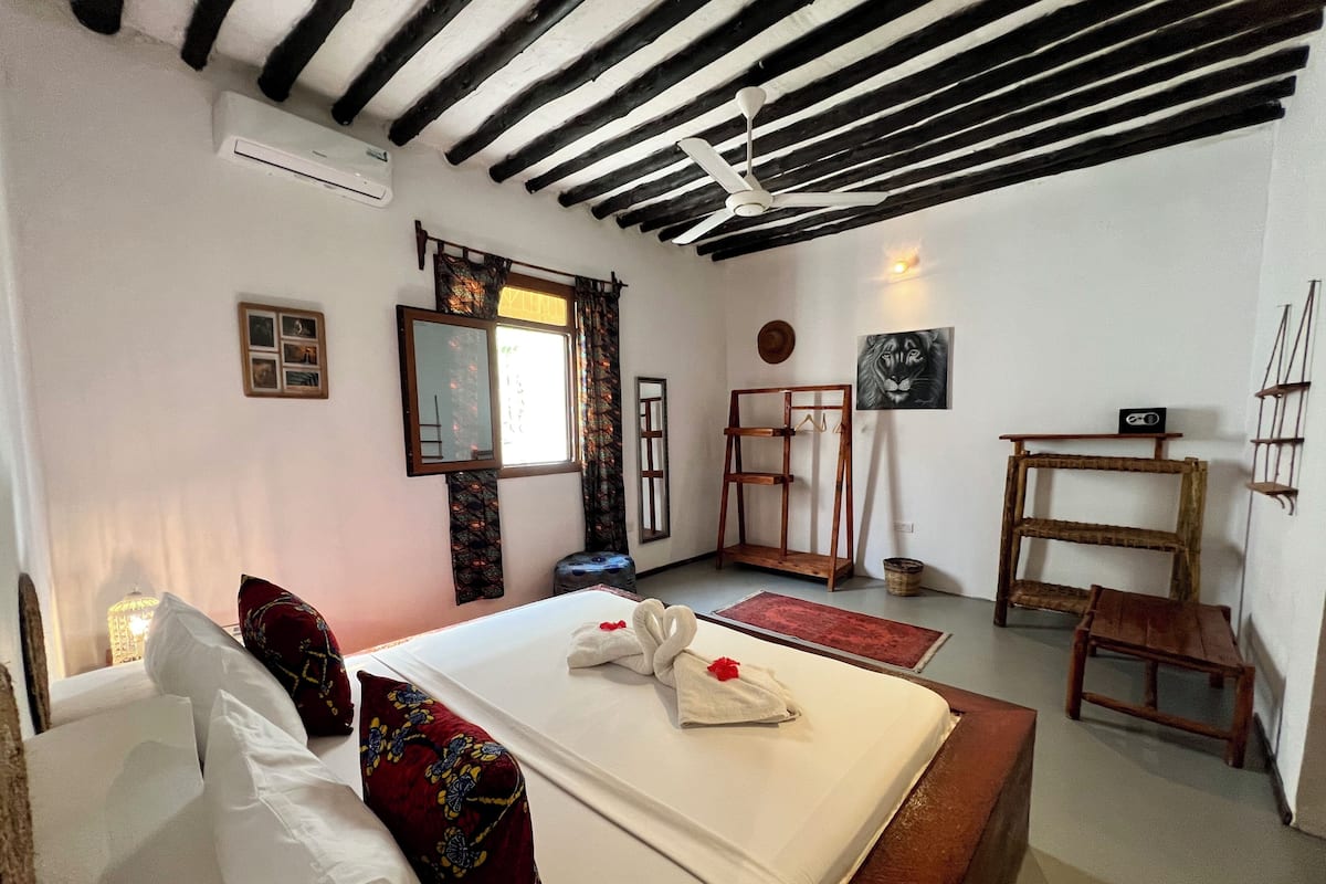 A bedroom with a bed, a wooden bench, a hanging chair, a mirror, a window with curtains, a wall-mounted shelf, and a ceiling fan.