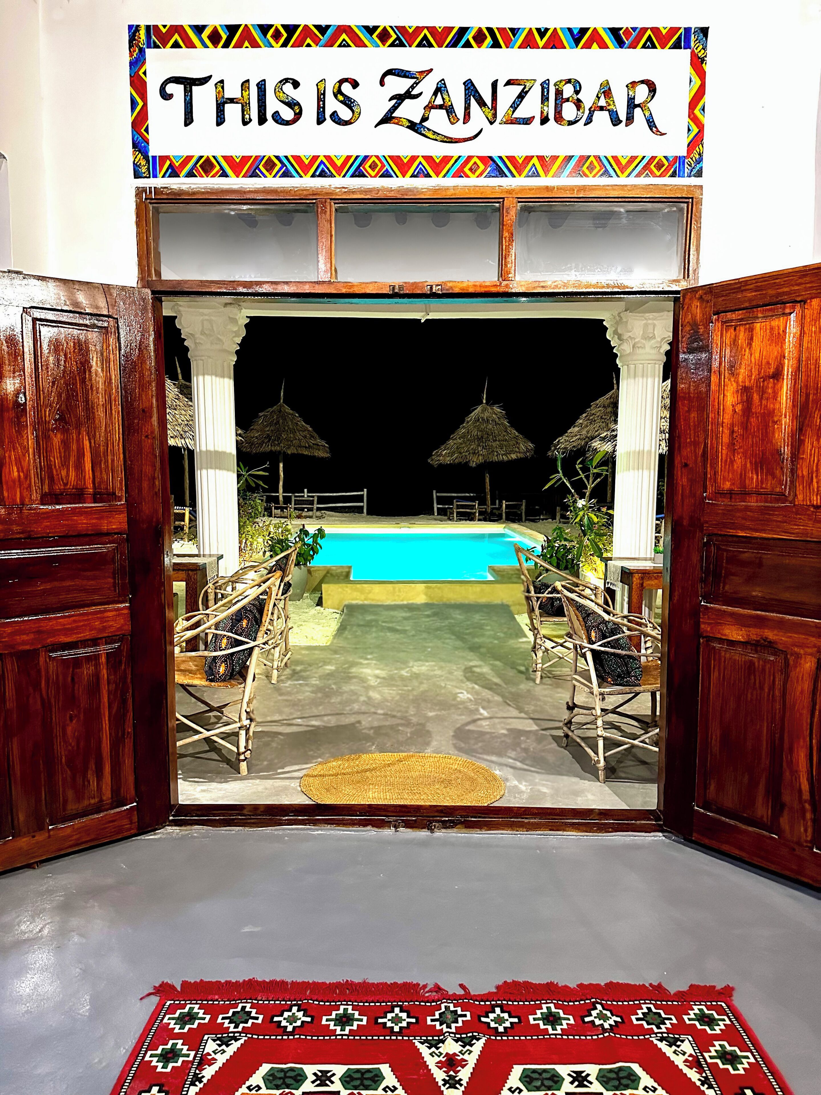 A wooden entrance with a sign reading 'THIS IS ZANZIBAR' leading to a pool area with a thatched umbrella and a woven chair.
