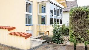 A residential building with a yellow facade, large windows, a small outdoor seating area, and a well-maintained garden with a hedge and rocks. - Awad's Yellow Apartment (Rust)