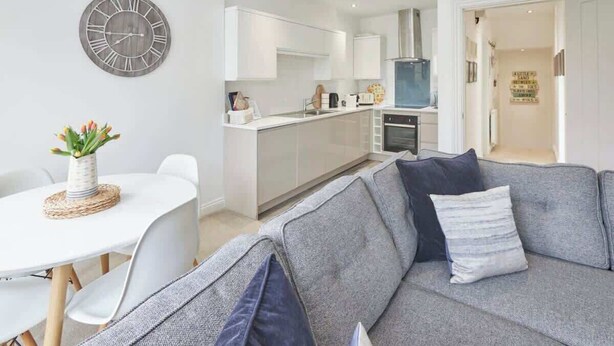 A modern living room with a grey sofa, white dining table, and a kitchen in the background. - Host & Stay | Number 2 at Pannett Apartments (Whitby)