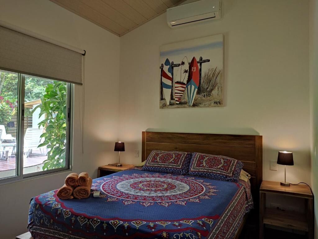 Ein Schlafzimmer mit einem großen Bett, zwei Nachttischen, einem Gemälde von Booten und einem Fenster mit Blick auf Grünflächen.
