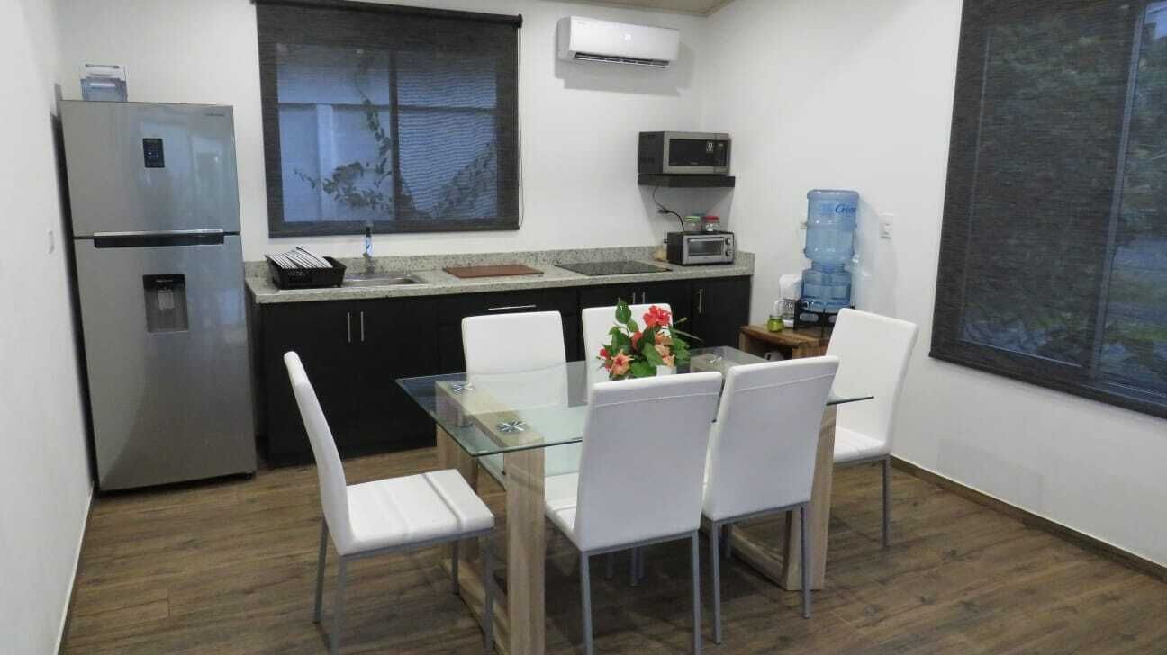 A compact kitchen with a refrigerator, microwave, and water dispenser. There is a dining table with chairs and a window with a view.