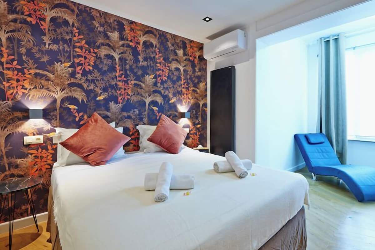 A hotel room with a large bed, two pillows, a bedside table, a wall-mounted air conditioner, a window with curtains, and a blue massage chair.