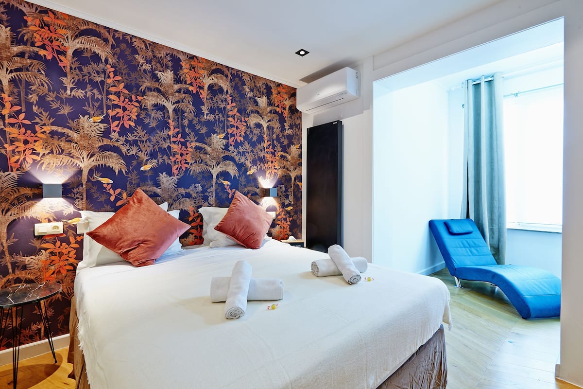 A hotel room with a bed, two pillows, a bedside table with a lamp, a blue lounge chair, and a large window.