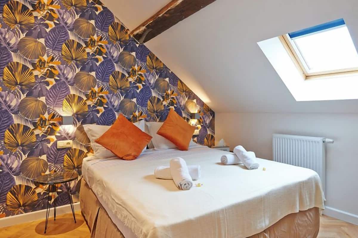 A bedroom with a sloped ceiling, a skylight, a bed with white linens, orange pillows, and a tropical leaf-patterned wallpaper.