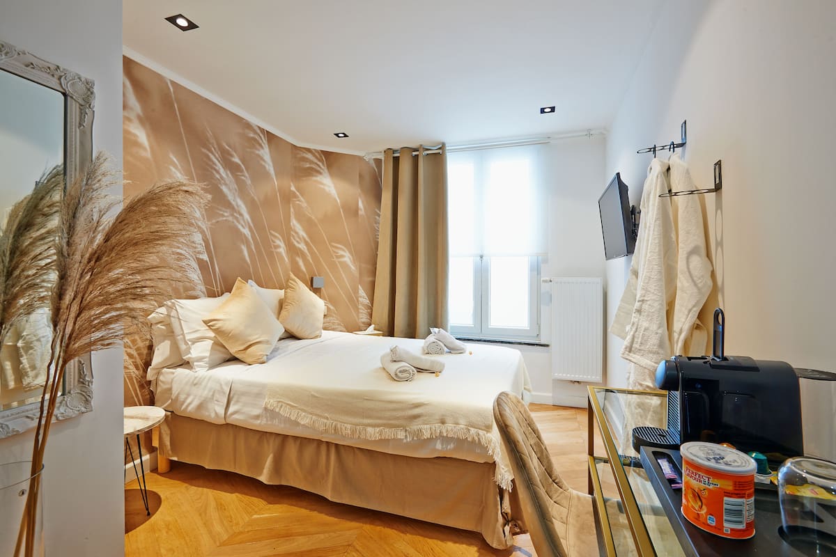 A hotel room with a bed, a glass table with a kettle, a coffee machine, and a window with curtains.