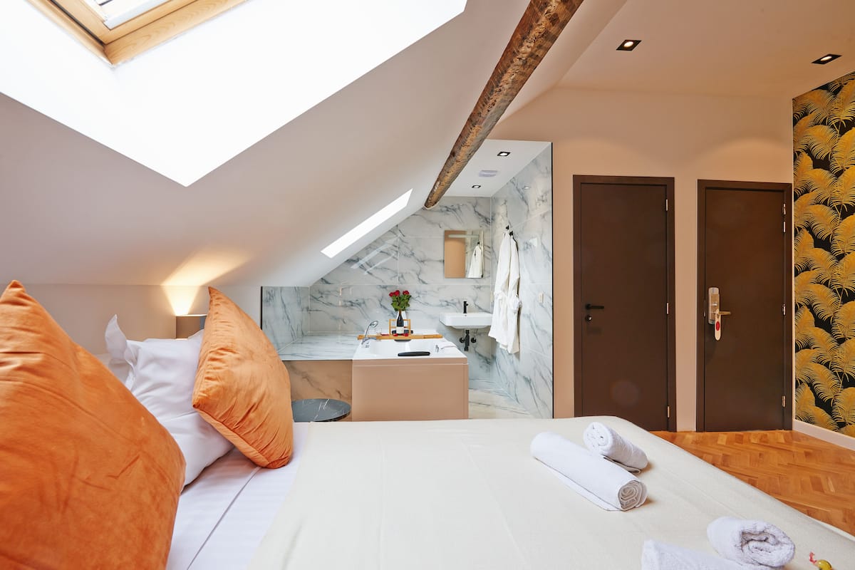 A modern hotel room with a skylight, a bed with orange pillows, a bedside table with a vase of flowers, a small bathroom with a marble sink, and two doors.