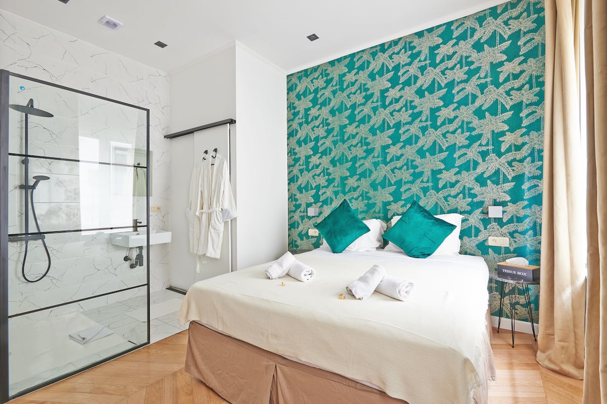 A hotel room with a glass-enclosed shower, a bed with neatly folded towels, a teal patterned wall, and a bedside table with a tissue box.