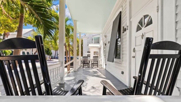A covered porch with two black chairs, a white door, and a palm tree in the background. - Mesa House Dos - One block from Duval St (Key West)
